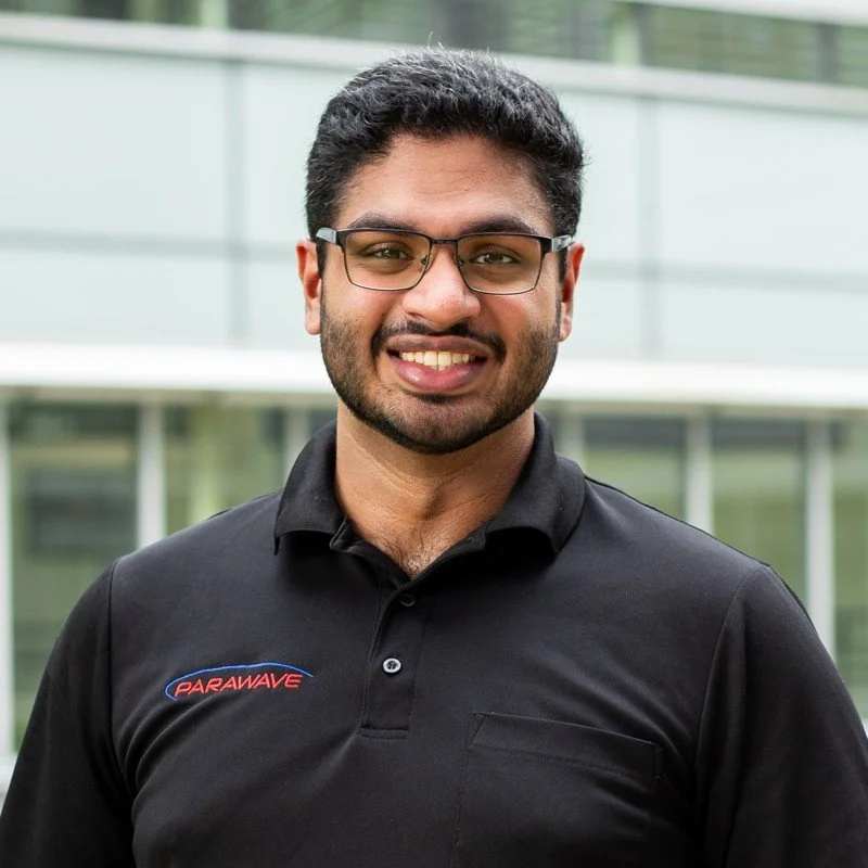 A smiling man wearing glasses and a black polo shirt with 'Parawave' logo, standing outdoors in front of a modern building.
