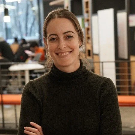 A woman with brown hair wearing a black turtleneck, smiling, in a coffee shop with large windows and people in the background.
