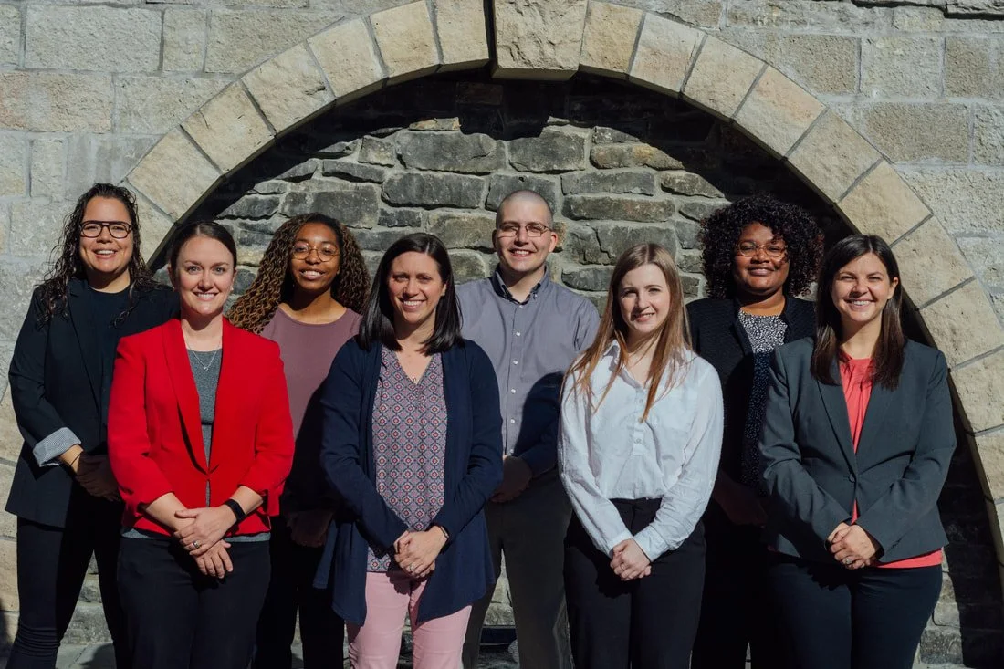 Group of nine diverse professionals standing outdoors in front of a stone archway, smiling.