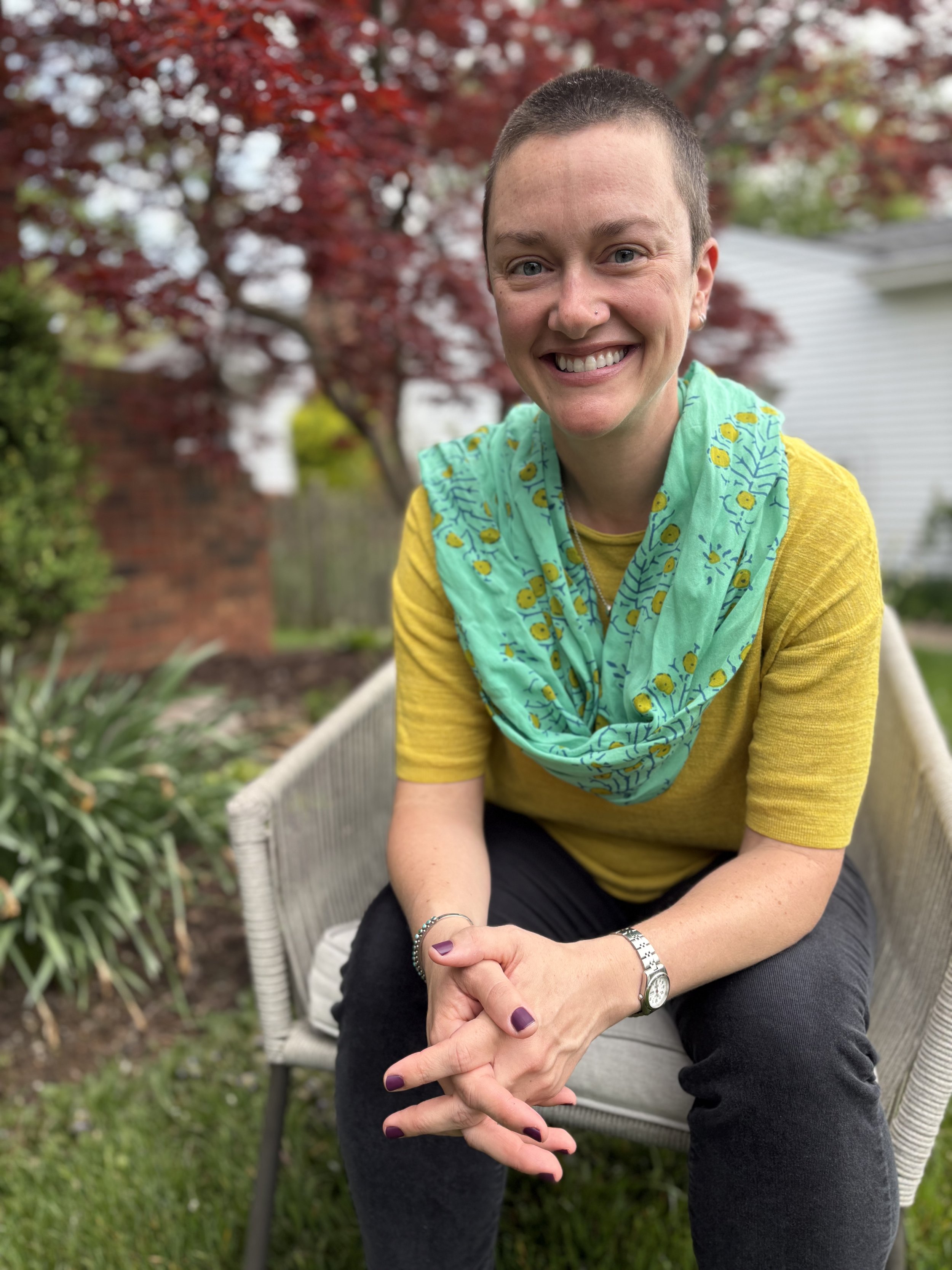 A woman with a short haircut and a nose piercing, smiling, sitting outdoors on a gray bench with a metal frame, with a red-leafed tree, green shrubs, and a house in the background. She is wearing a yellow top, a green scarf with yellow flowers, and has a silver watch and bracelet on her left wrist, and painted purple nails.