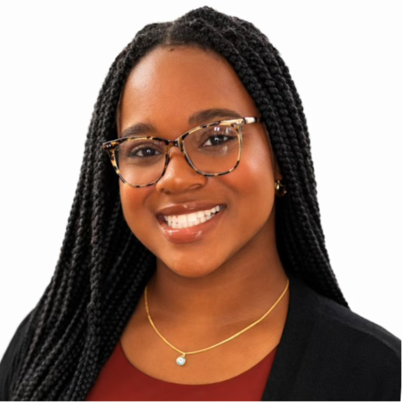 A woman with long braided hair wearing glasses, smiling, and dressed in black and burgundy with jewelry.