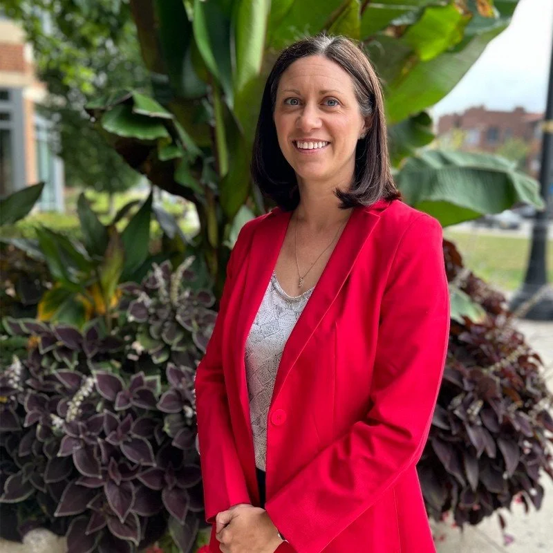 A woman with shoulder-length brown hair smiling in front of lush greenery, wearing a red blazer over a light-colored top.
