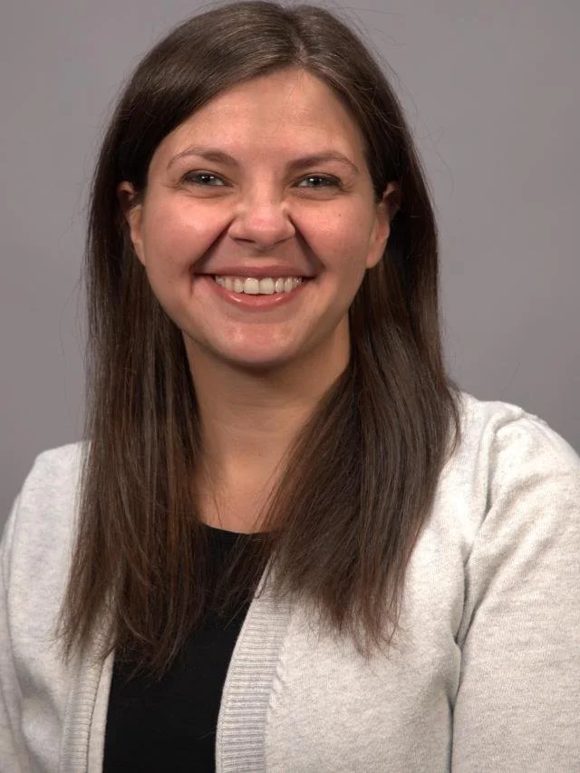 A young woman with long brown hair, smiling, wearing a light gray cardigan over a black top, against a plain gray background.