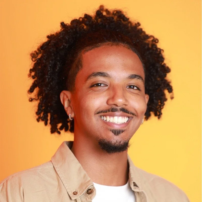 Portrait of a smiling man with curly hair, earrings, and a goatee, wearing a beige shirt against a yellow background.