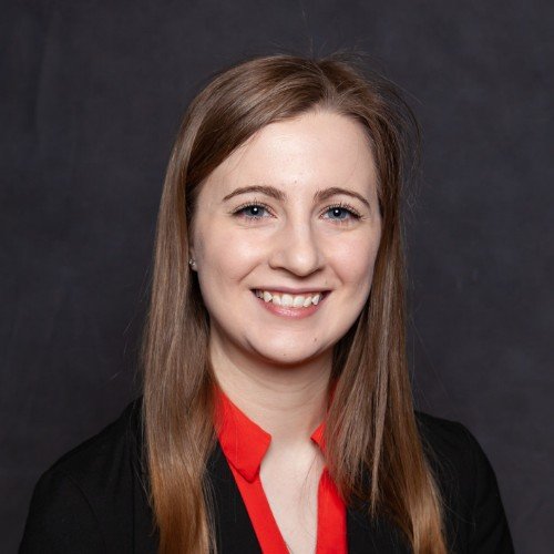 A young woman with long, straight, light brown hair, smiling, wearing a black blazer and a red blouse, against a dark background.