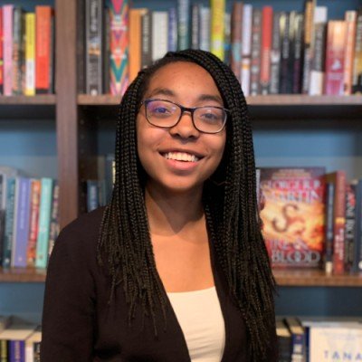 A young woman with glasses and braided hair smiling in front of a bookshelf filled with colorful books.