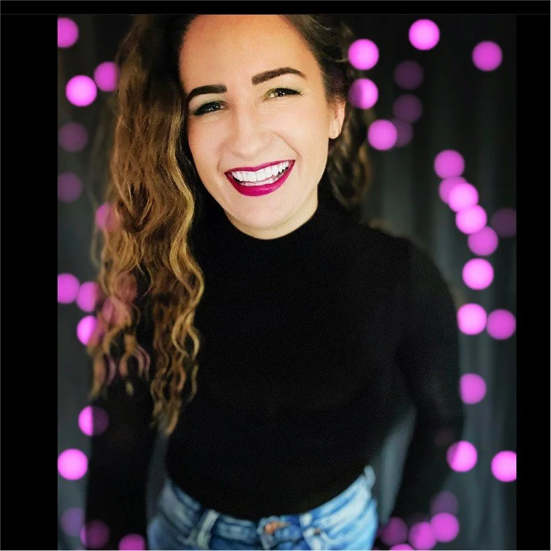 A woman smiling, wearing a black turtleneck and jeans, with long, wavy hair, standing against a backdrop decorated with purple bokeh lights.