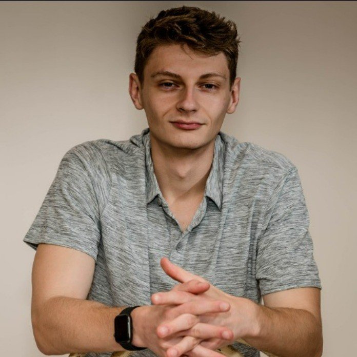A young man with short, wavy brown hair, wearing a light gray polo shirt and a black smartwatch, is sitting against a plain beige background, looking directly at the camera with a slight smile, hands clasped together on the table.