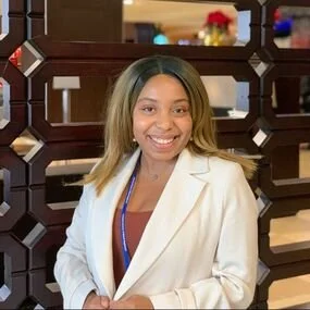 A young woman with shoulder-length hair wearing a white blazer and a lanyard, smiling and standing indoors in front of a decorative partition with a dark wood frame.