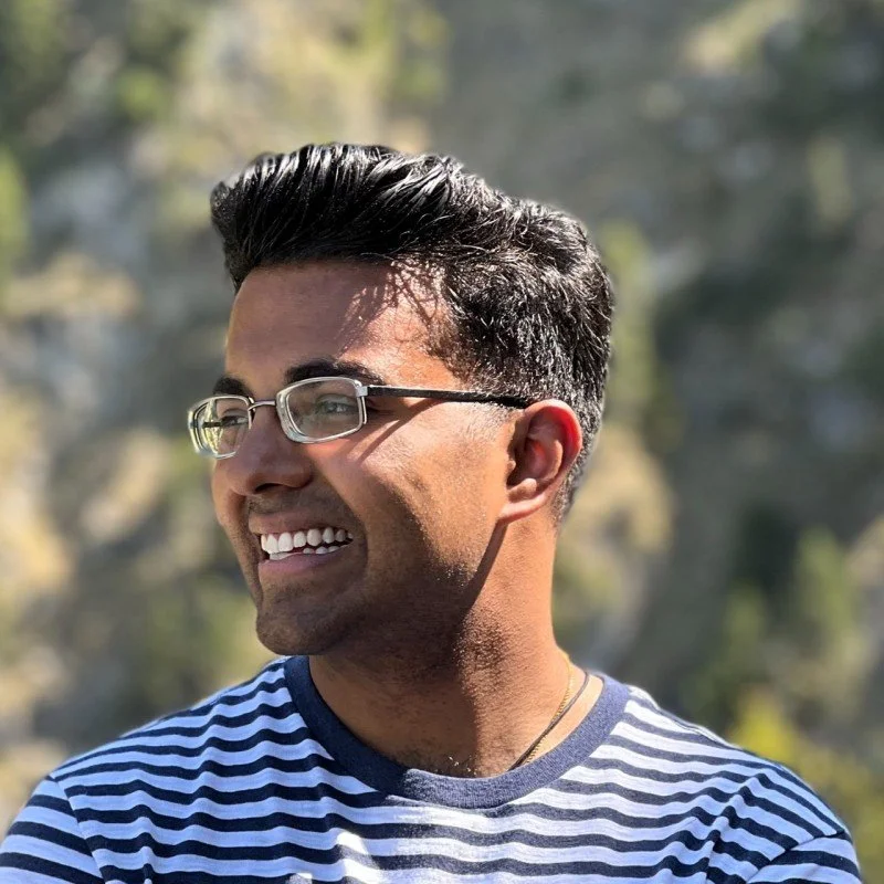 A smiling man with glasses and dark hair outside, wearing a striped shirt, with blurred trees in the background.