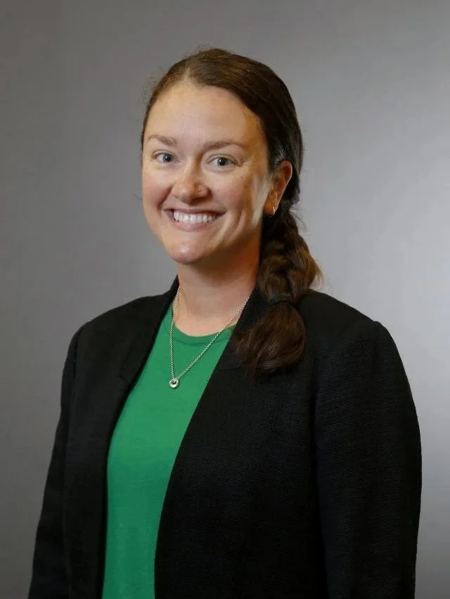 Portrait of a woman with long brown hair in a side braid, wearing a black blazer and a green top, smiling against a plain gray background.