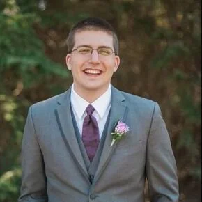 A young man in a gray suit and purple tie, smiling outdoors with a boutonniere on his lapel.