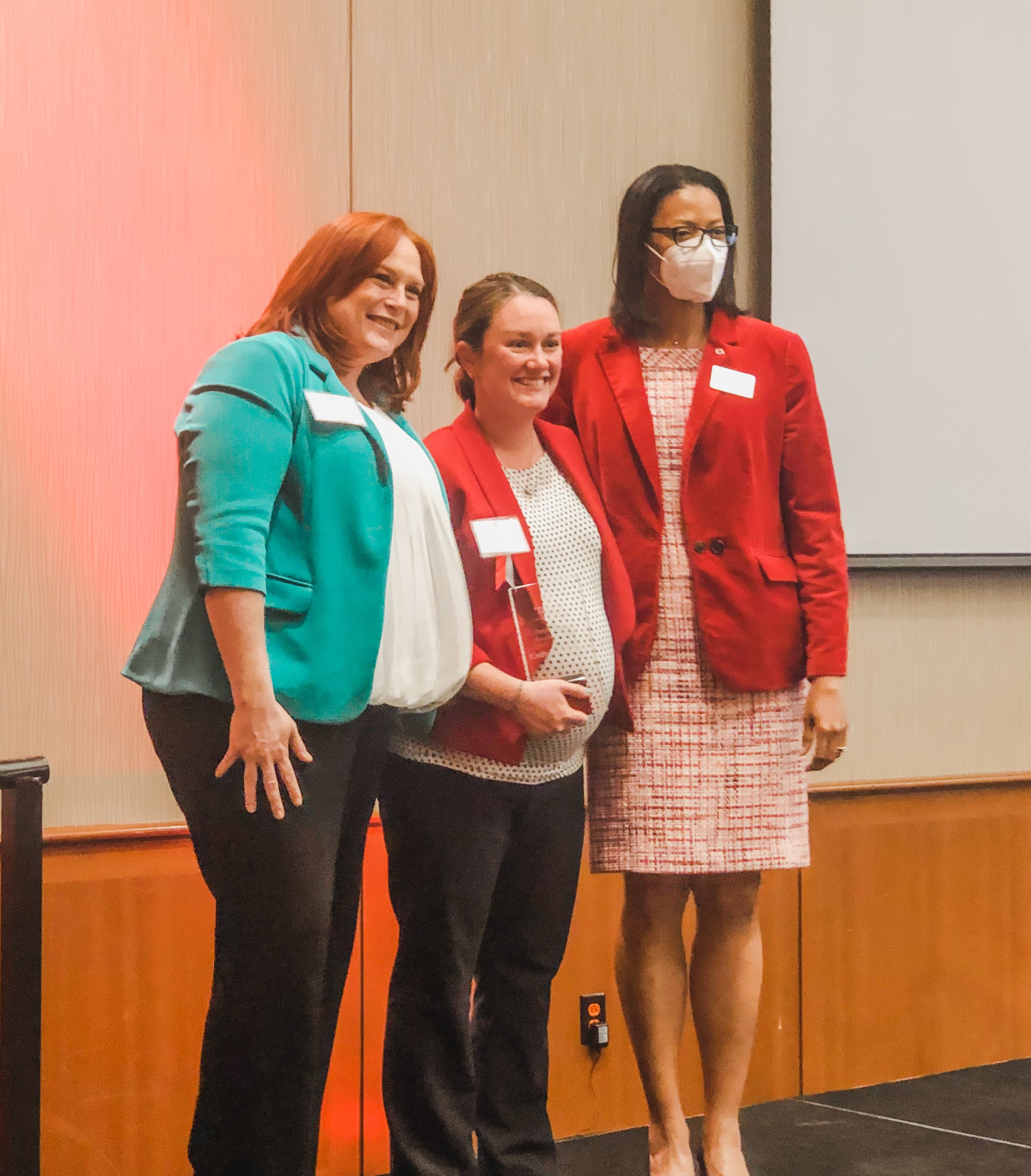 Three women standing side by side indoors in front of a wood panel wall, smiling and posing for the camera. The woman on the left has red hair, is wearing a teal blazer and black pants. The woman in the middle has light brown hair, is wearing a red blazer over a polka dot blouse. The woman on the right has dark hair, glasses, a face mask, and is wearing a red blazer over a patterned dress.