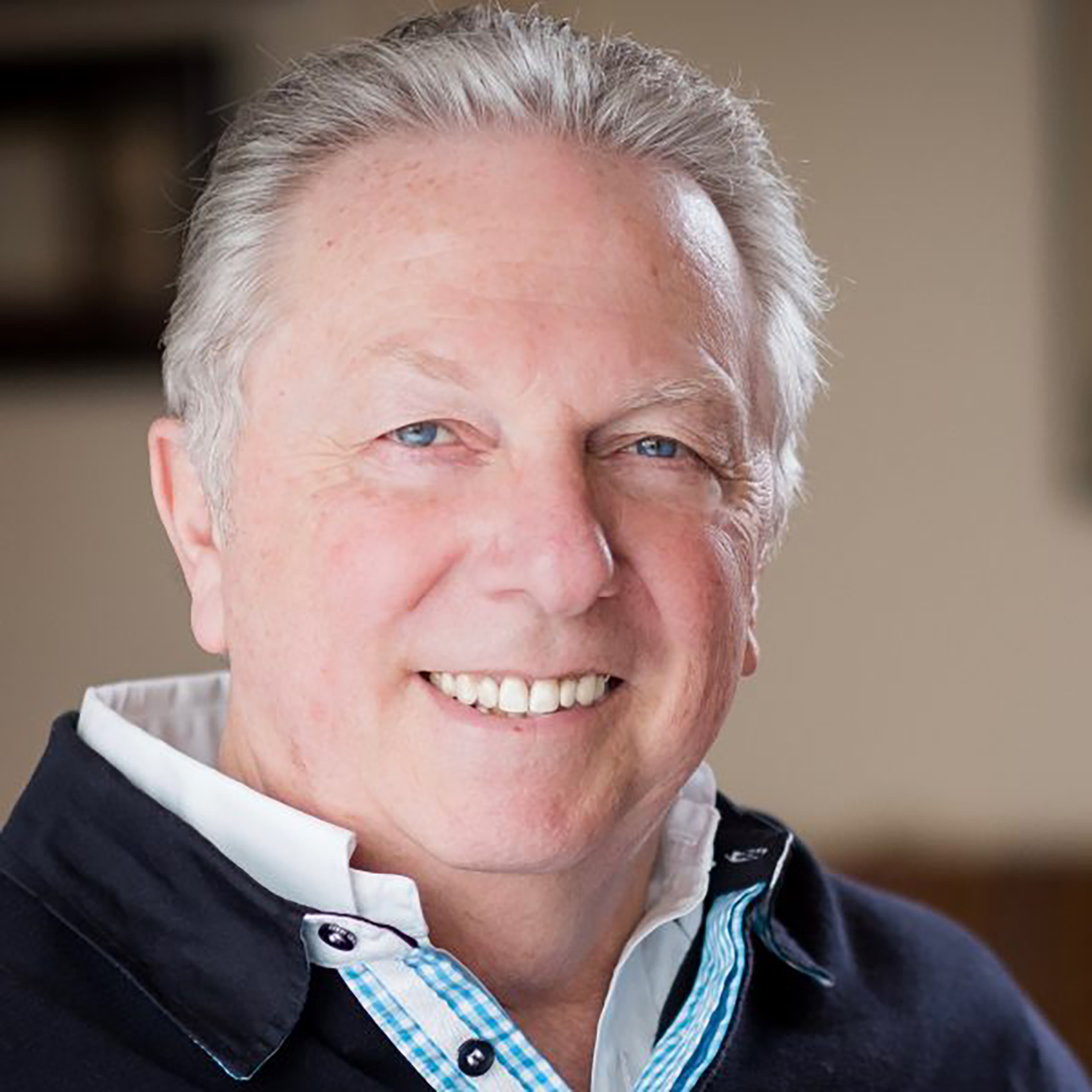 Close-up of John Halker with light gray hair, blue eyes, and fair skin, wearing a black jacket over a collared shirt with a checkered pattern.
