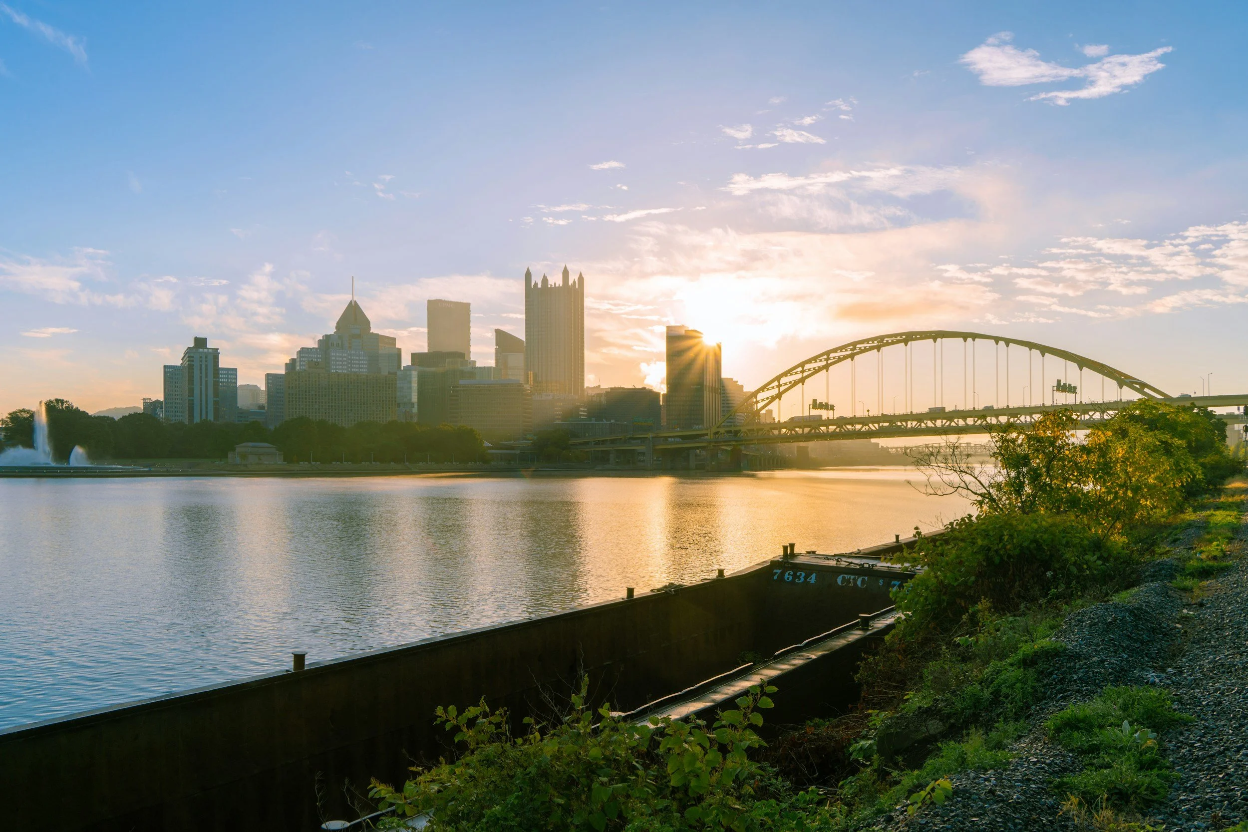 City skyline during sunset with high-rise buildings, a river in the foreground, and a bridge arching over the river, with green trees along the riverbank.