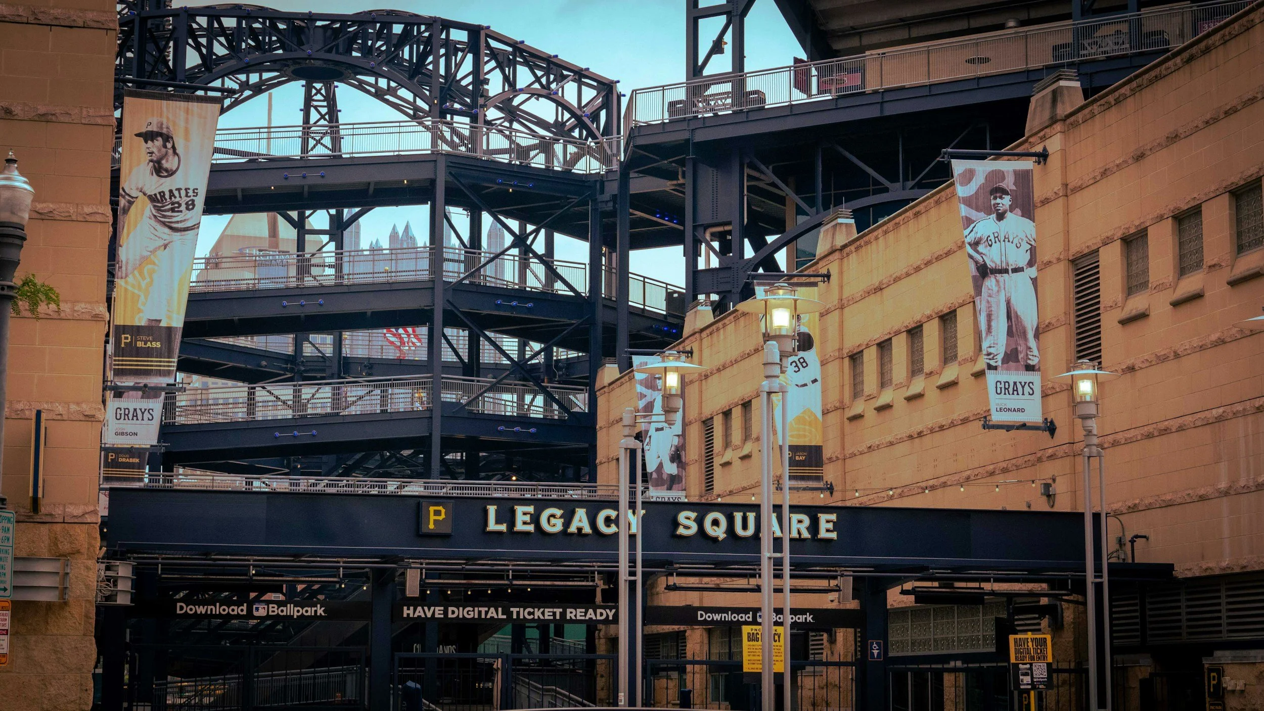 A view of the entrance to the Pirate's baseball stadium, Legacy Square, with banners of historical baseball players and a structure with multiple walkways and stairs. The background shows a city skyline with tall buildings.