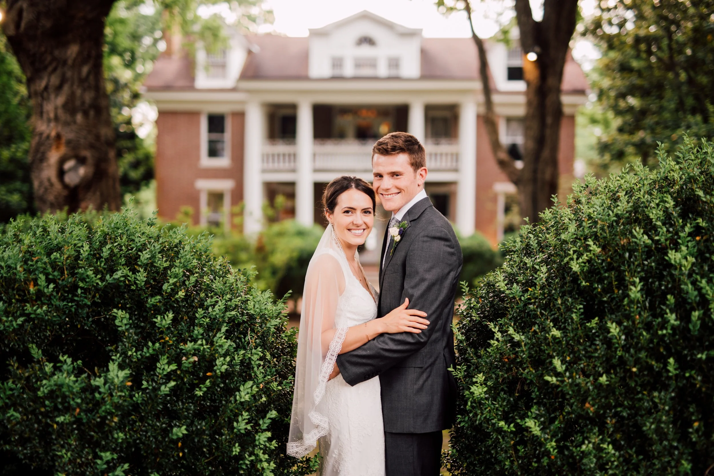 A bride and groom smiling and embracing in front of a large house with a porch, surrounded by greenery and trees.