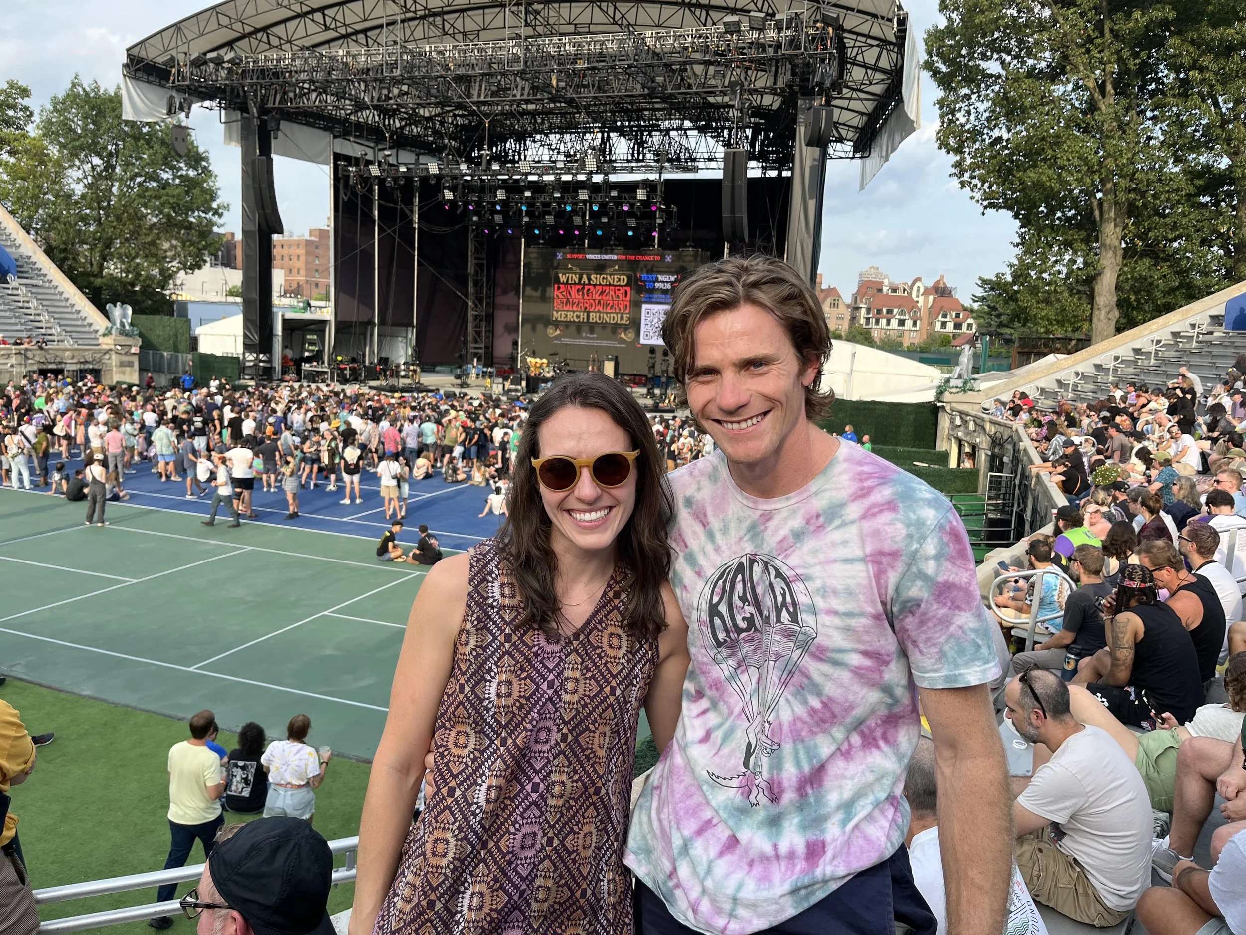 Two smiling people, a woman dentist pretty with sunglasses and a man in a tie-dye shirt, at an outdoor concert at Forest Hills King Gizzard and the Lizard Wizard with a large stage and many seated and standing attendees in the background.