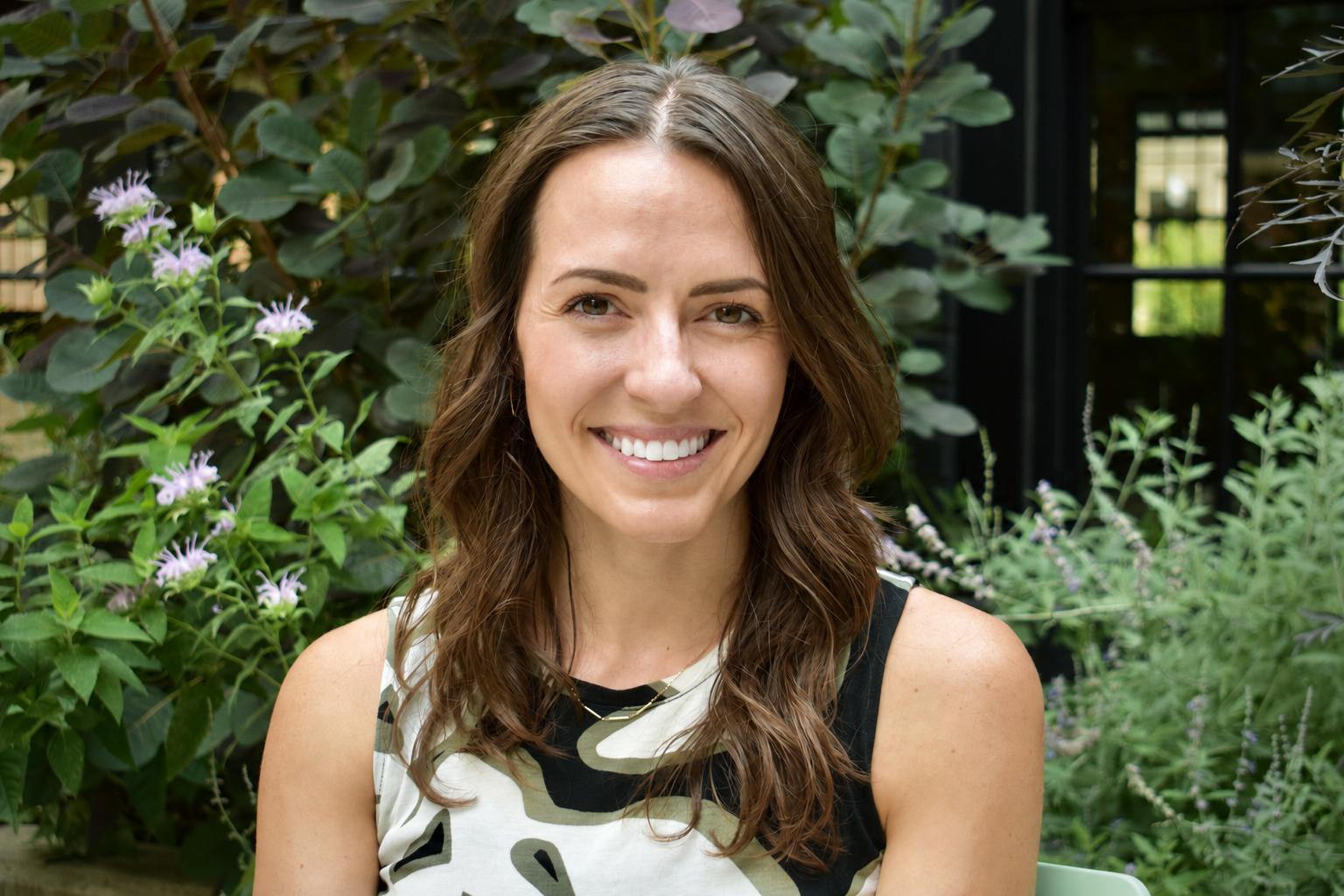 A woman dentist pretty with wavy brown hair and a bright smile sitting outdoors in front of lush green plants and purple flowers.