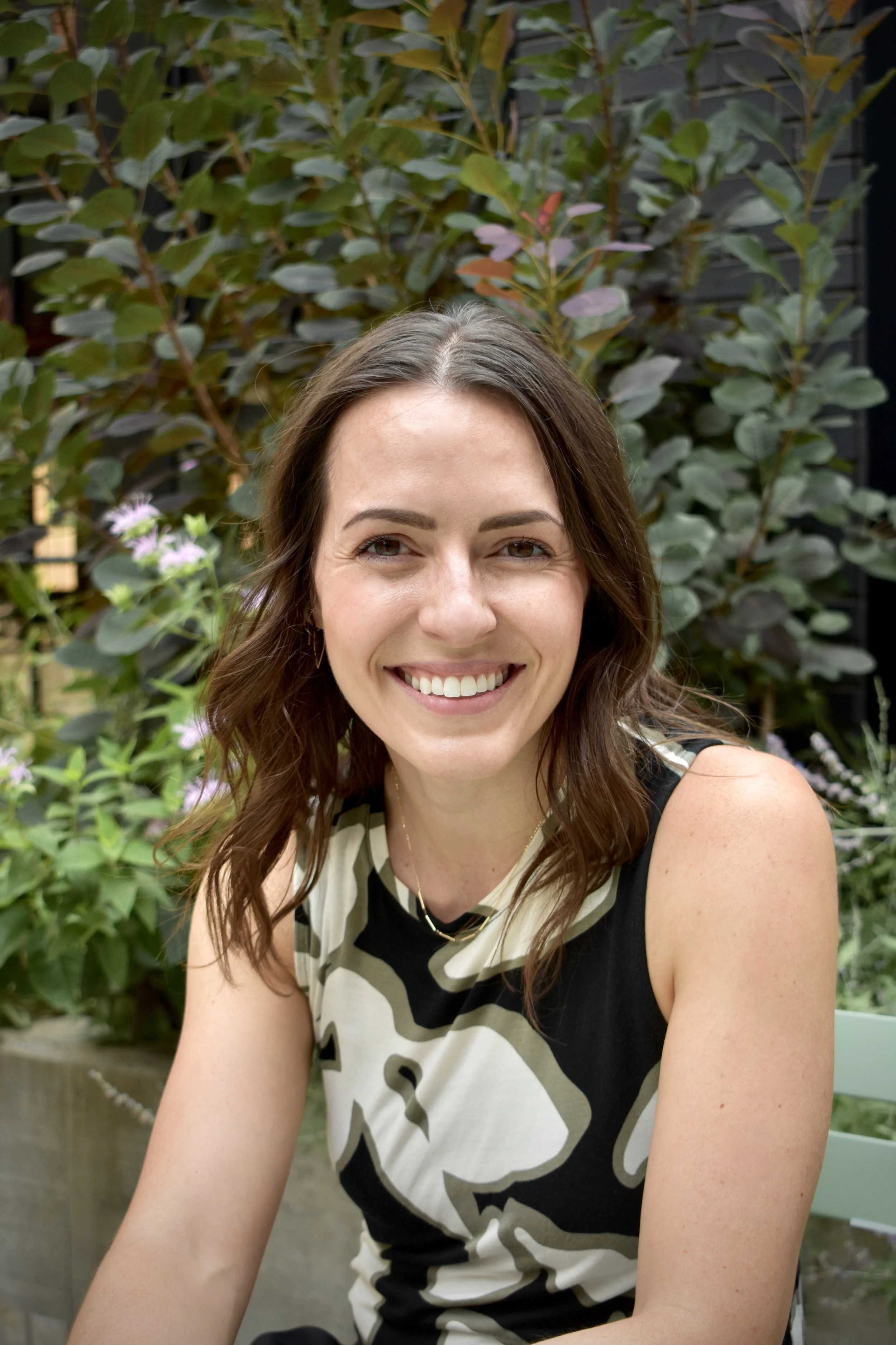 Close-up of a woman dentist pretty with brown hair, smiling, wearing a patterned sleeveless top, sitting outdoors with green plants and a dark wall in the background.