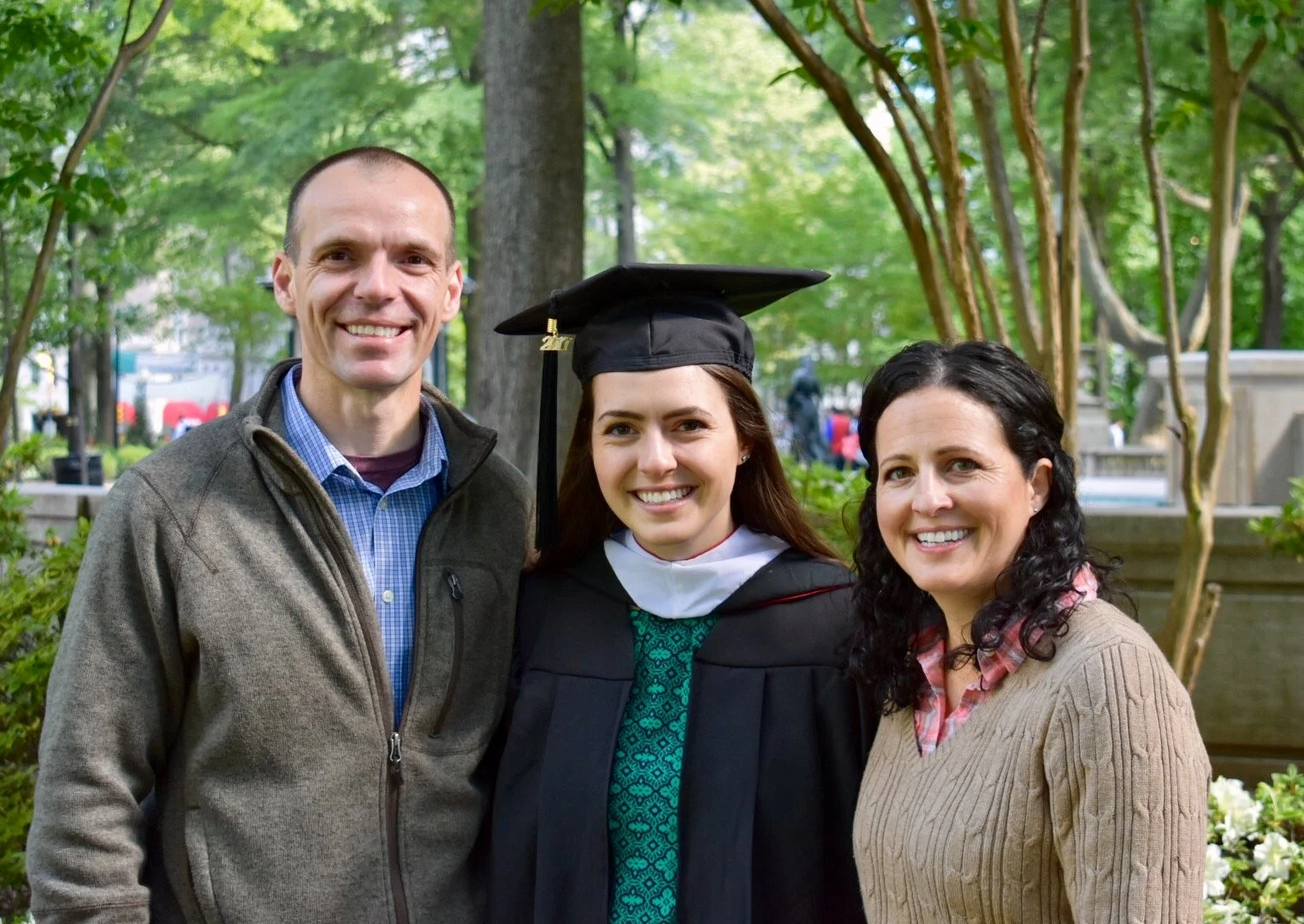 A young woman in a graduation cap and gown smiling between two adults, outdoors with trees and greenery in the background.