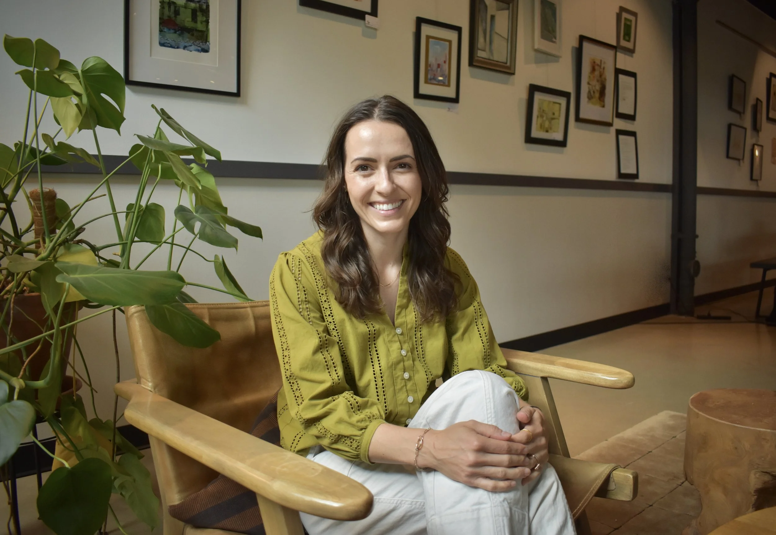A woman dentist pretty with wavy brown hair, wearing a yellow blouse, sitting on a wooden chair with a smile in a gallery or cafe with framed artwork on the wall and greenery nearby.