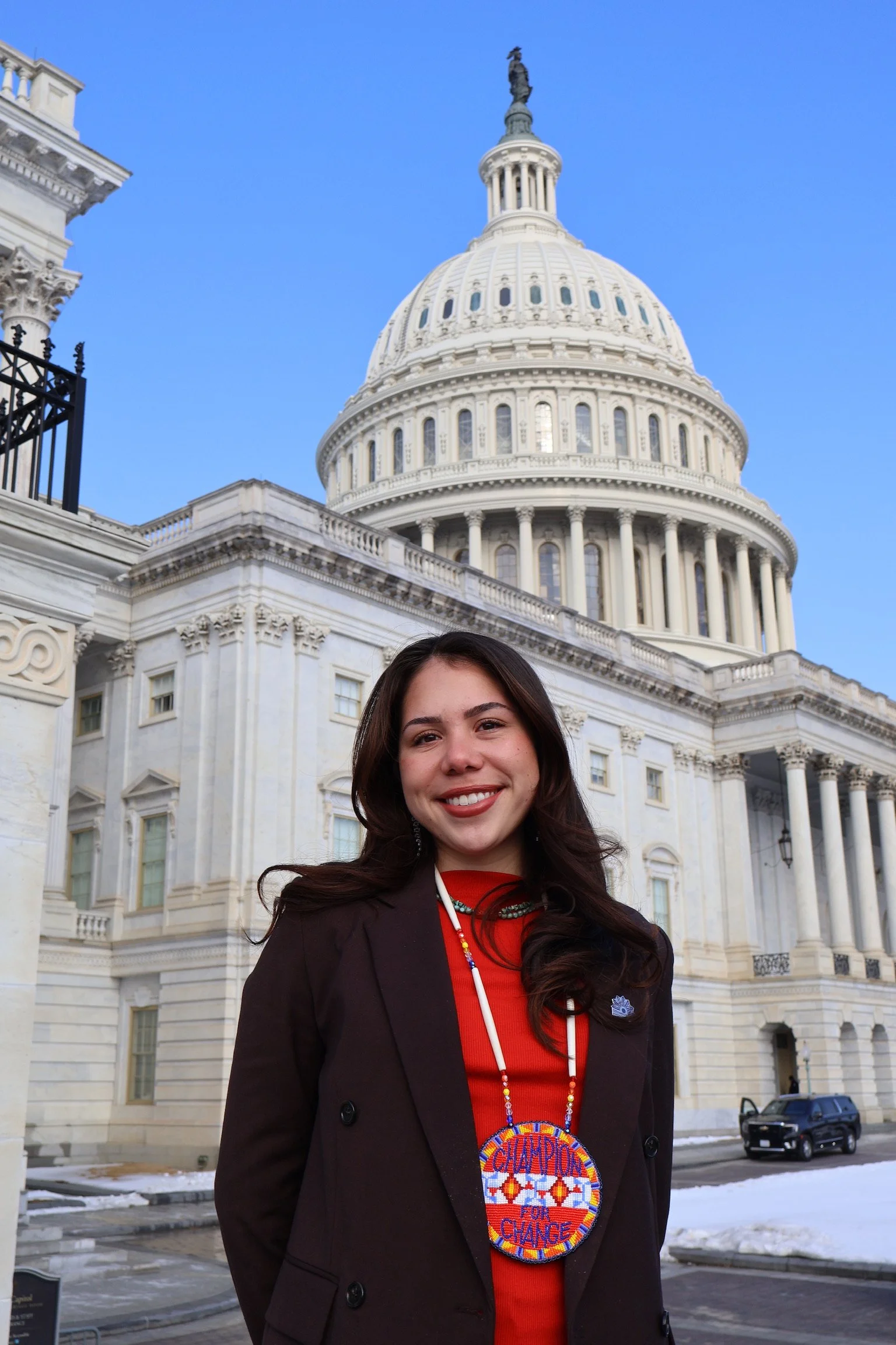 A smiling woman with long dark hair standing in front of the United States Capitol building. She is wearing a dark blazer over a red top, and a large colorful badge that reads 'Champions for Change'. There is snow on the ground and a black car parked behind her.