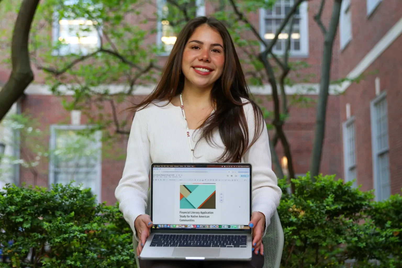 A young woman holding a laptop outdoors with a project on the screen titled 'Financial Literacy Application Study for Native American Communities'. She has long brown hair, is smiling, and is wearing a white sweater. She is sitting in front of greenery and a brick building with multiple windows.