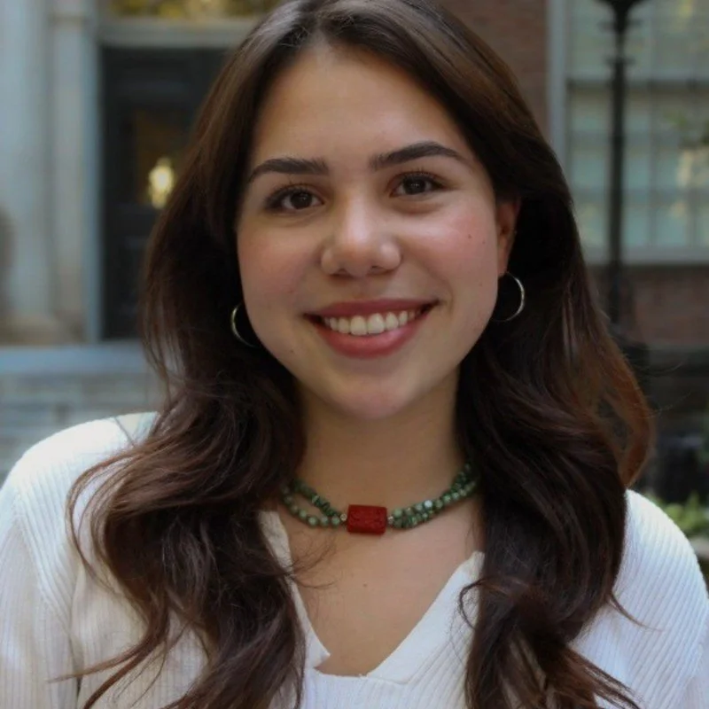 A young woman with long brown hair, smiling, wearing earrings and a beaded necklace, outdoors in front of a building.
