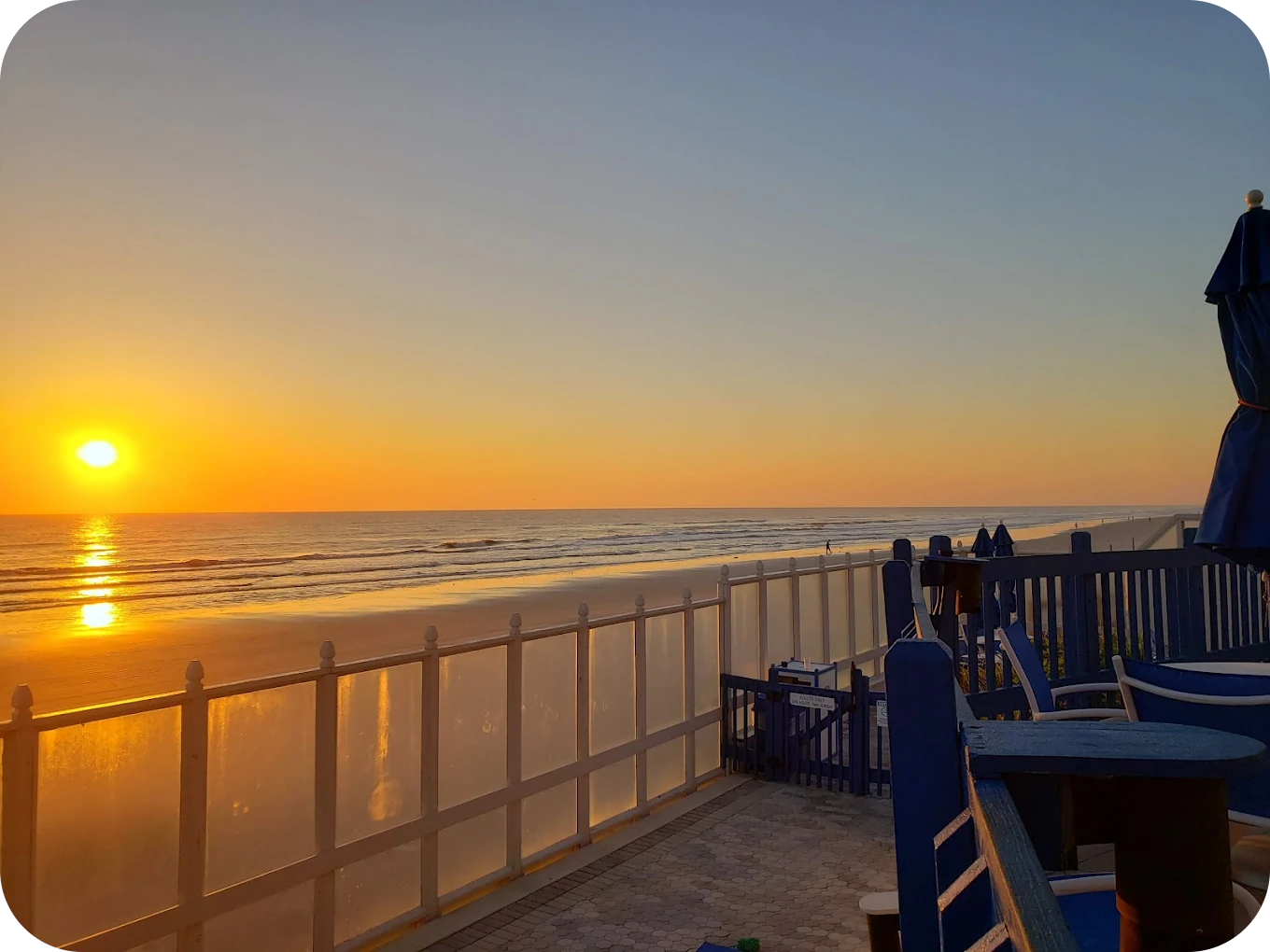 Sunset over the ocean viewed from a beachfront patio with blue chairs and umbrellas, and a white fence.