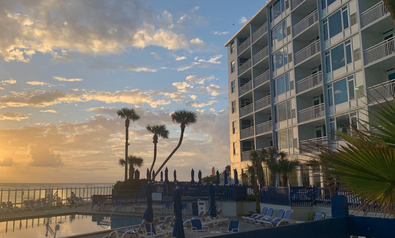 A hotel pool area at sunset with lounge chairs, umbrellas, and palm trees in front of a large multi-story building by the ocean, with a partly cloudy sky.
