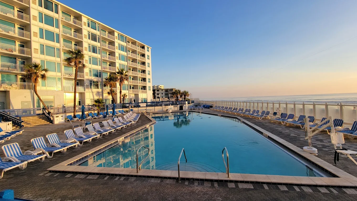 Empty swimming pool at a beachfront hotel with blue and white striped lounge chairs, palm trees, and the ocean in the background during sunset.