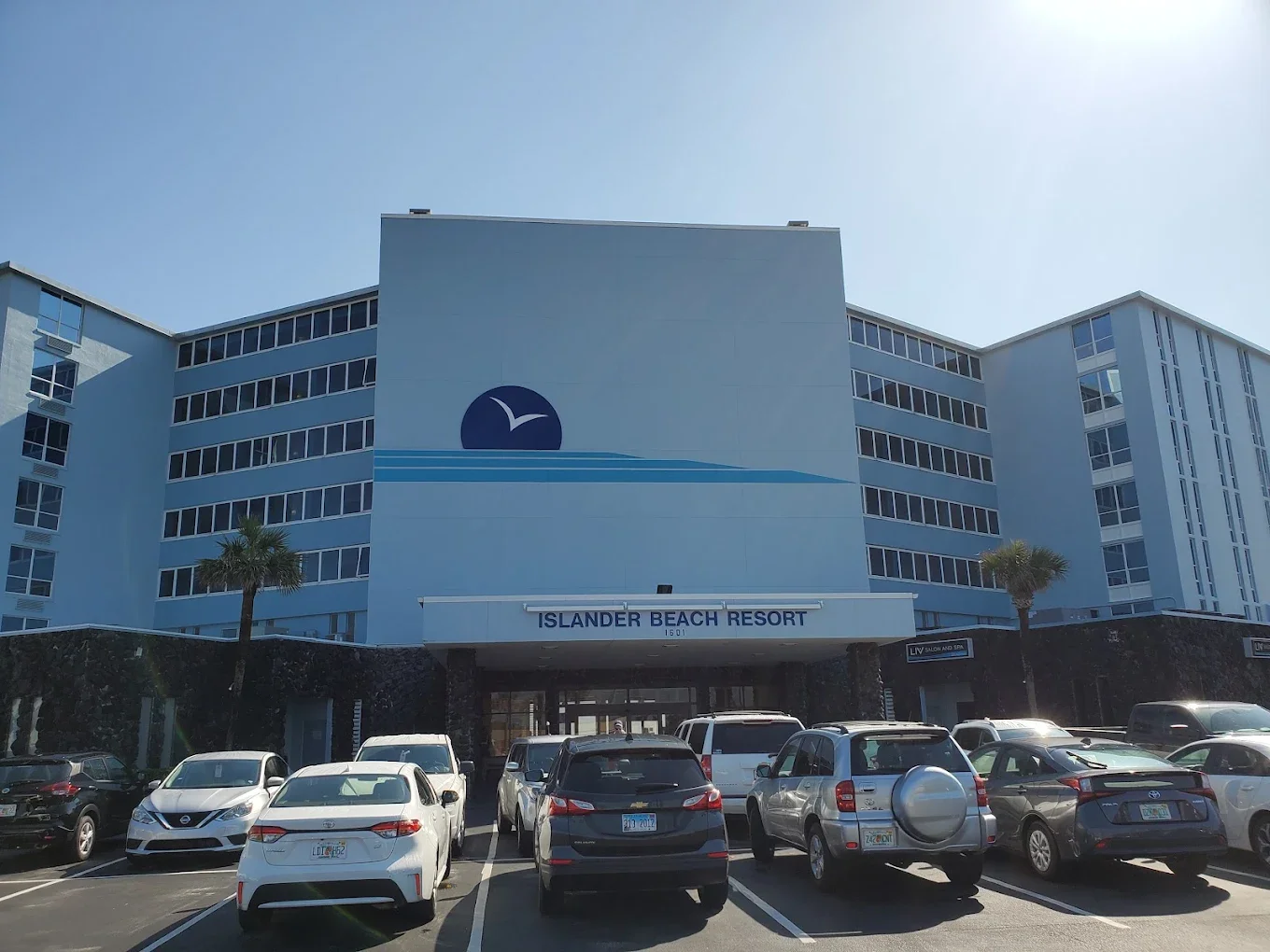 Blue multi-story hotel building with the sign 'Islander Beach Resort' above the entrance, with parked cars and palm trees in the foreground.