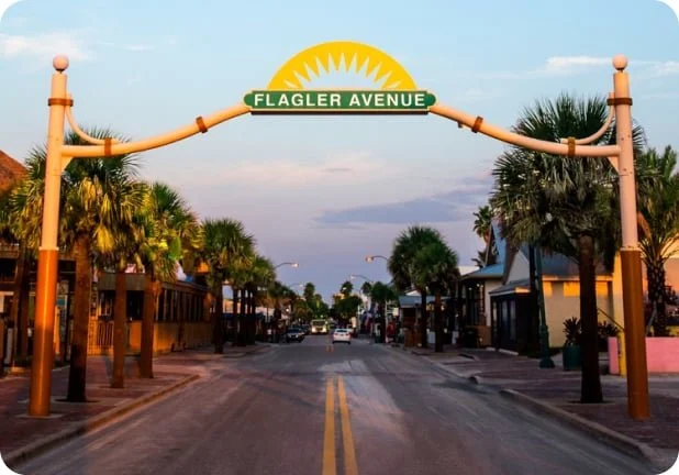 Entrance arch with a yellow sun design and green sign that reads 'Flagler Avenue,' over a small town street lined with palm trees.