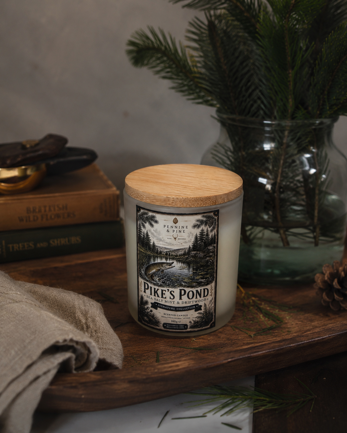 A scented candle labeled 'Pike's Pond' with pine illustrations, placed on a wooden tray next to a glass vase with pine branches, and books about British wildflowers and trees.