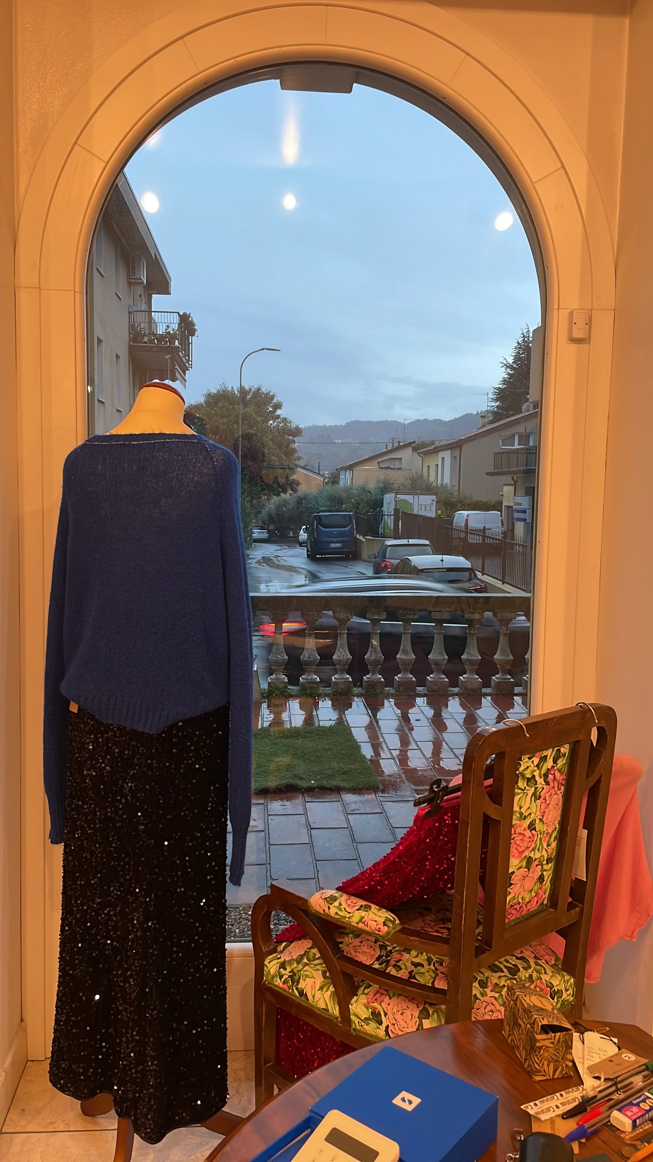 View through an open door to a rainy street with parked cars, residential buildings, and trees in the background. Inside, a mannequin dressed in a blue sweater and a black sequin skirt is positioned near the door. There are chairs with floral upholstery, a table with various items including a tissue box, writing utensils, and a remote control.