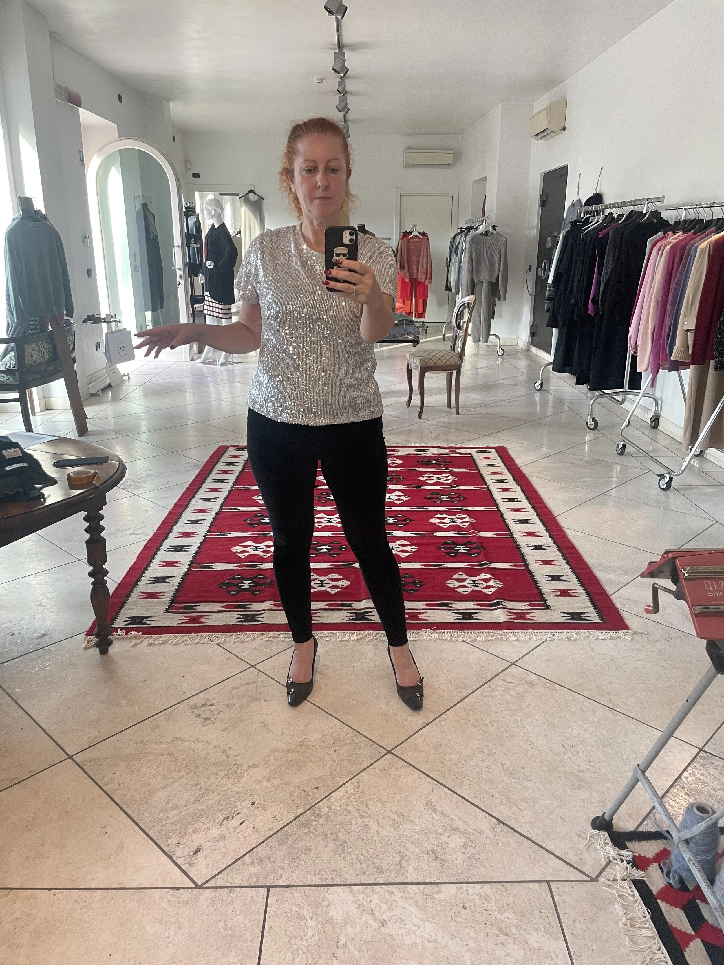 A woman with red hair taking a mirror selfie inside a clothing store. She is wearing a sparkly silver top, black pants, and black high heels. The store has clothing racks with various garments, a red and black patterned rug, a small sofa, a table with accessories, and a mannequin in the background.