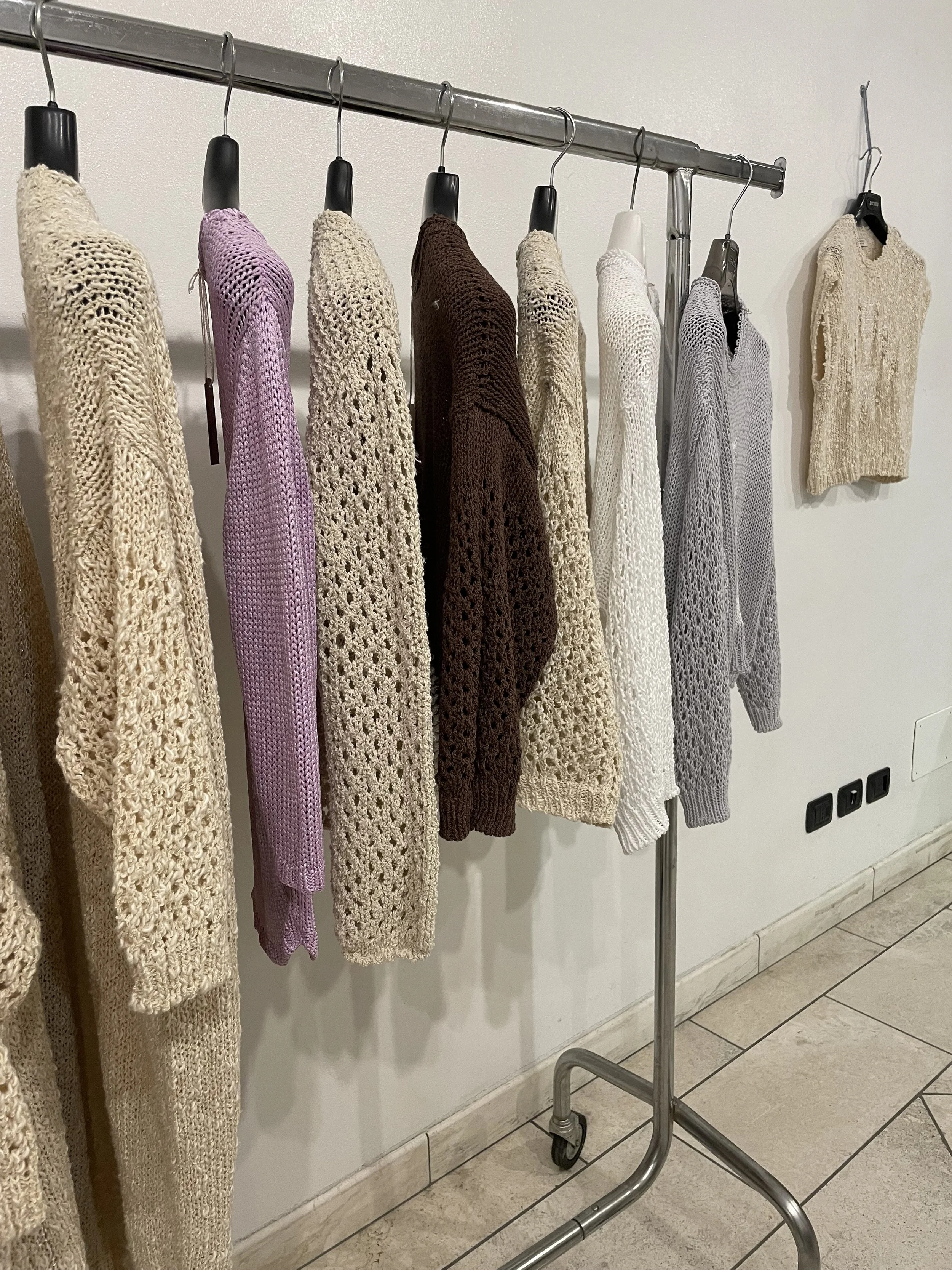 A metal clothing rack on wheels with various knitted sweaters hanging on it, against a white wall with electrical outlets and a tiled floor.