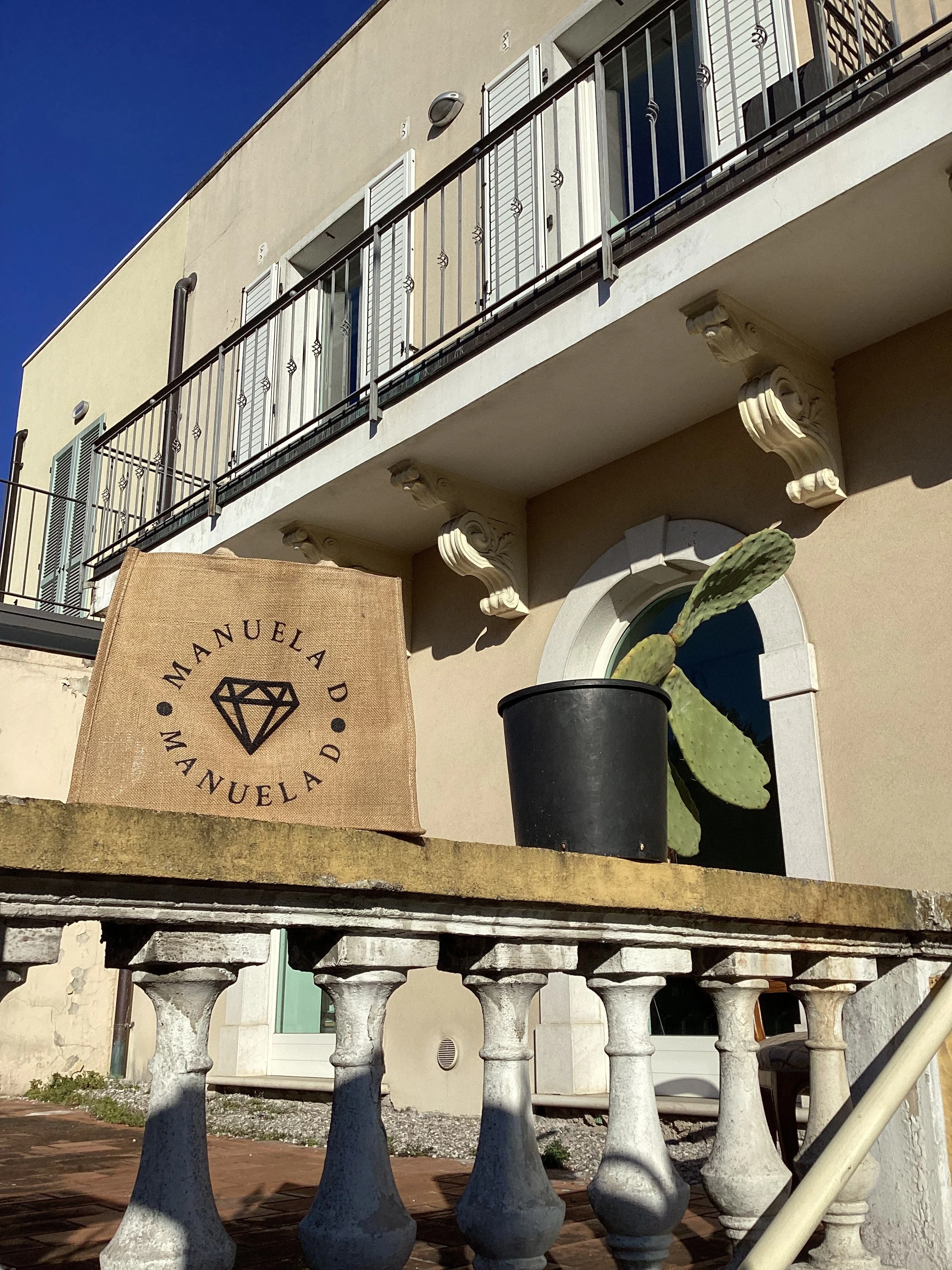 View of a building's upper level with a balcony and white shutters, a cactus in a black pot on a ledge, and a decorative stone balustrade below. There is a sign with a diamond logo and the words 'MANUELA D' partially visible on the left side.
