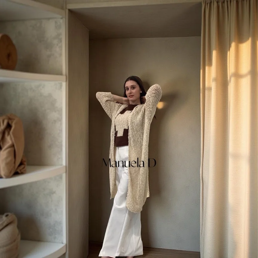 A woman with her arms raised behind her head stands in a small alcove with beige walls, wearing a beige cardigan, brown top, and white pants, beside a window with beige curtains.