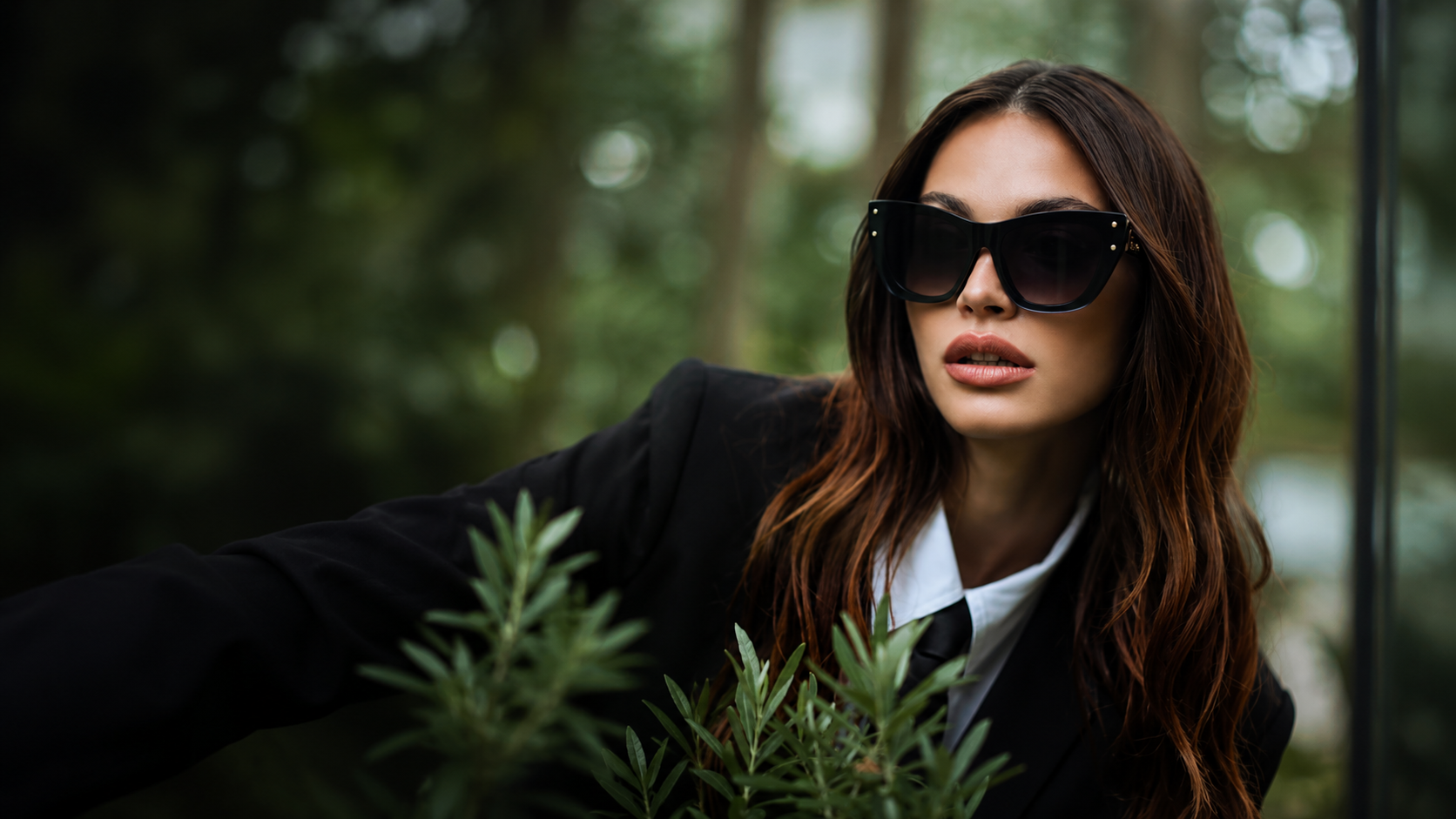 A woman with long brown hair wearing black sunglasses, a black blazer, and a white shirt, reaching out to touch green plants outdoors with blurred trees in the background.