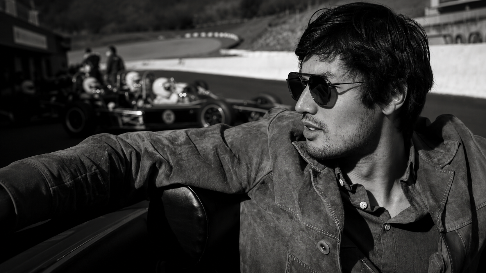 A man with dark hair and sunglasses, wearing a leather jacket, sitting in a car at a race track with vintage race cars in the background.