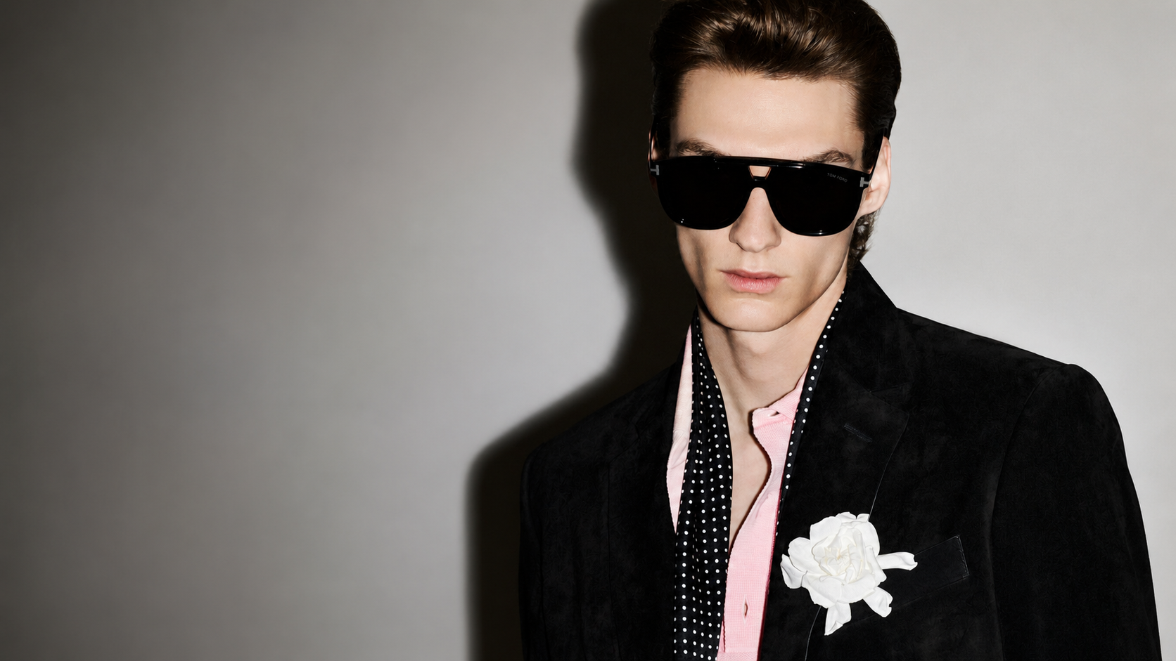 A young man with brown hair wearing large black sunglasses, a black blazer, pink shirt, black and white polka dot scarf, and a white flower pin on the blazer, standing against a gray background.