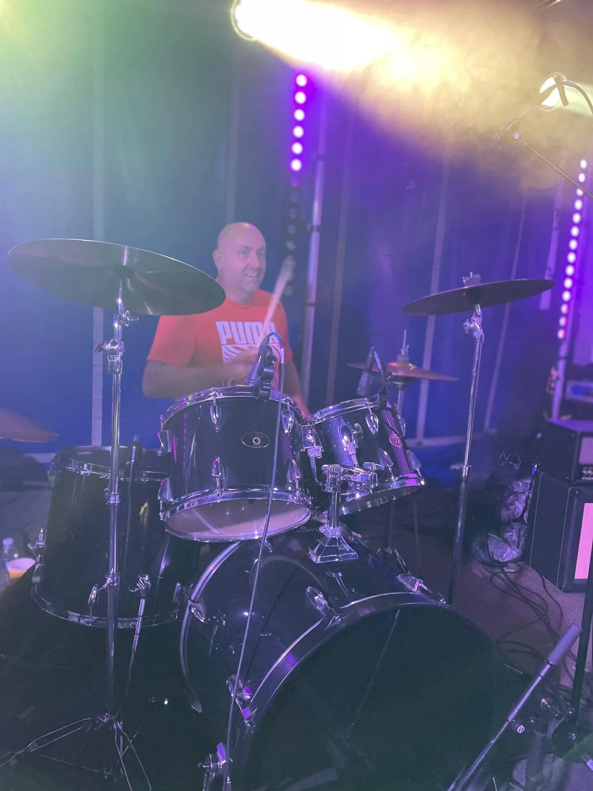 A man playing a drum set on stage, wearing a red T-shirt with the word 'Puma' on it, surrounded by stage lighting and equipment.