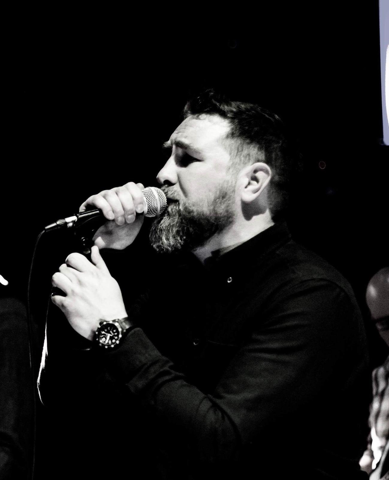 A man singing into a microphone during a performance, with a beard and wearing a watch, in a dark setting.