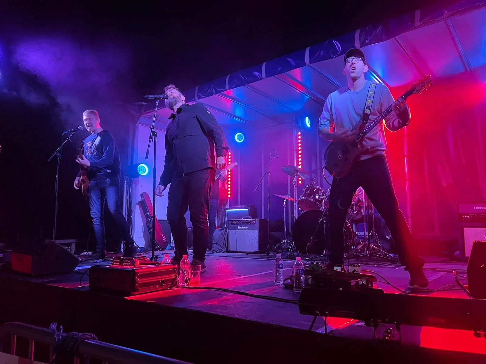 A band performing live on stage with colorful lighting, three musicians playing guitars and a drummer in the background.