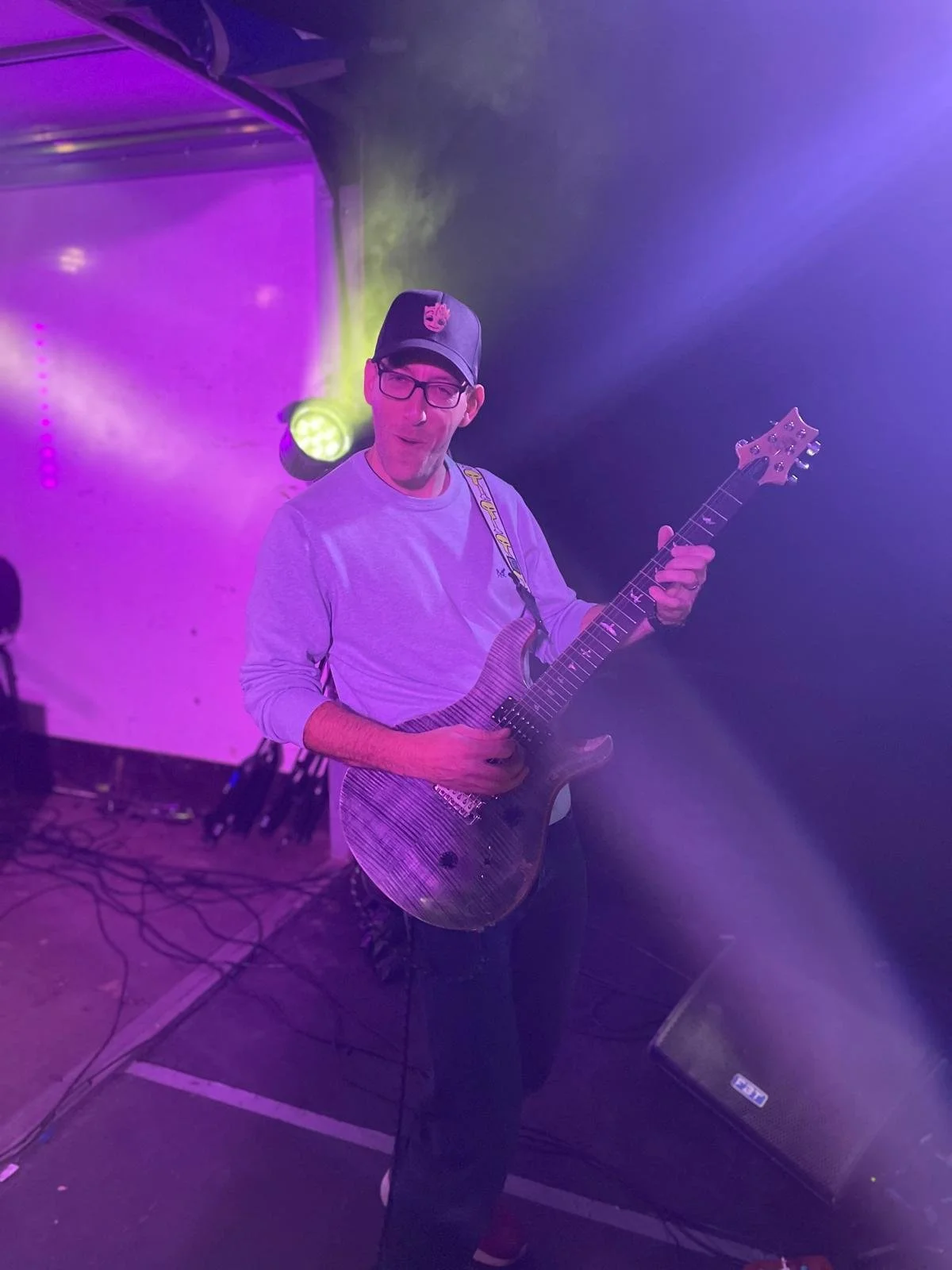 A man playing an electric guitar on stage, wearing glasses, a gray sweatshirt, a baseball cap with a pin, under purple and yellow stage lights.