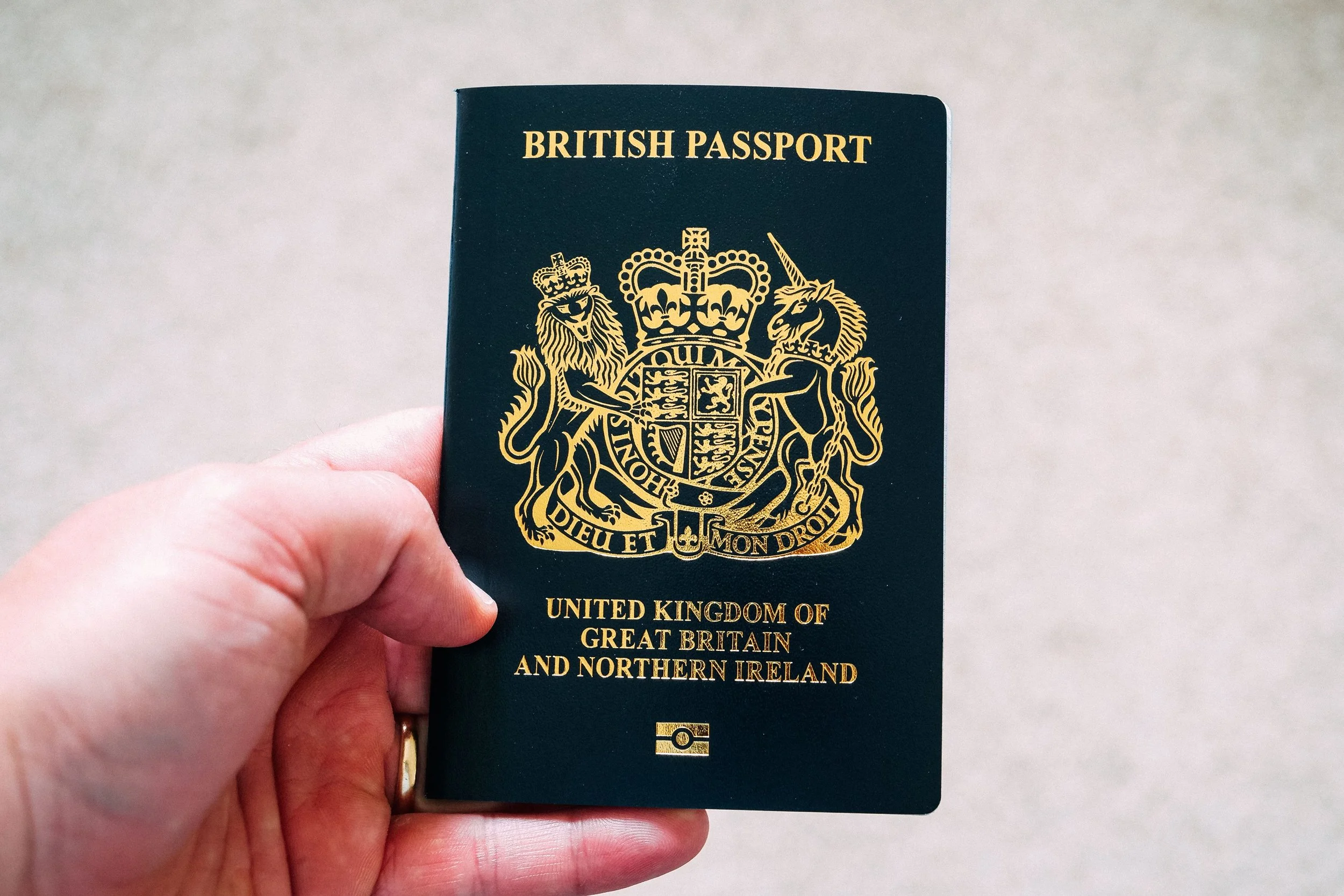 A hand holding a British passport with a gold emblem on the cover, showing the royal coat of arms of the United Kingdom.