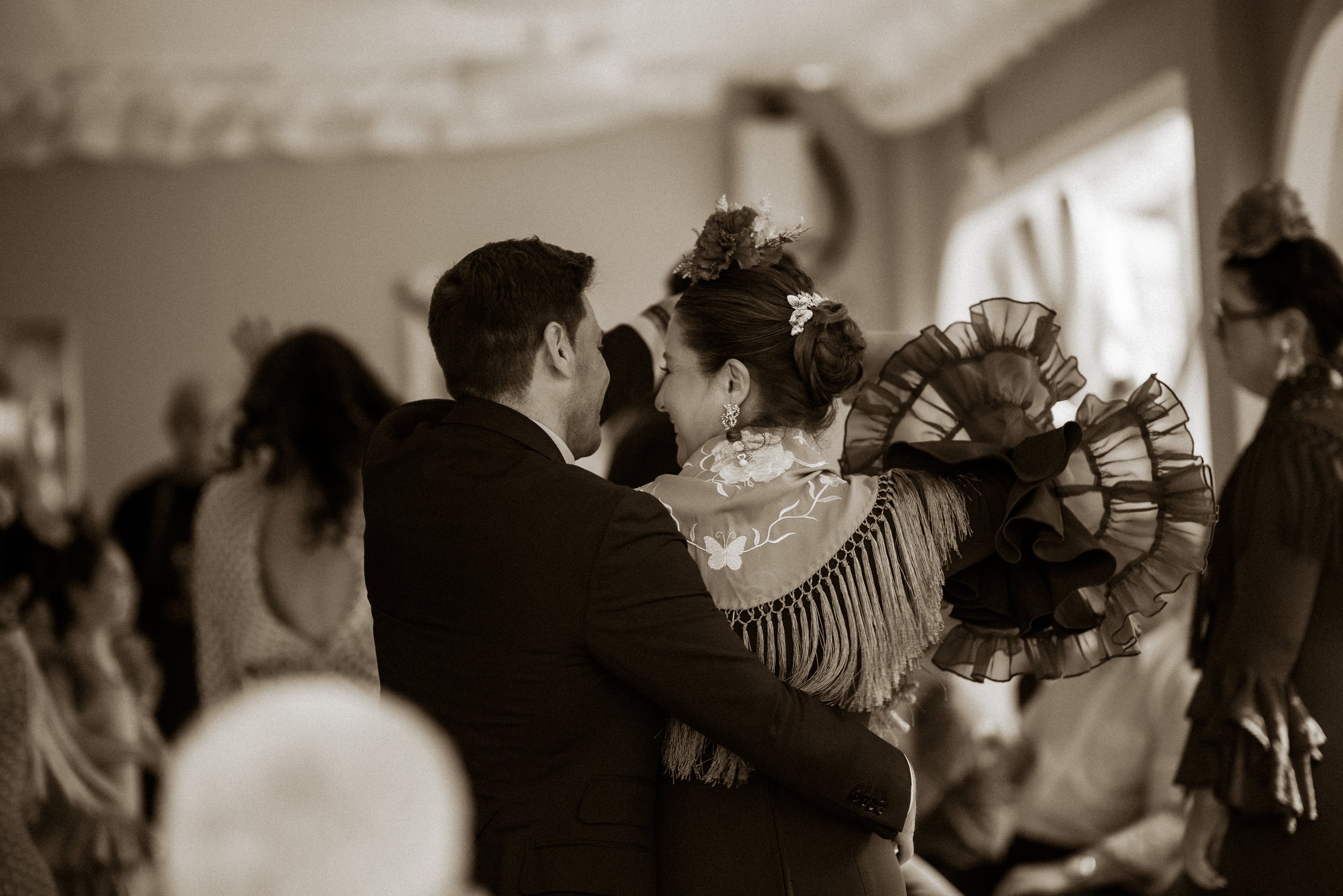 A couple dancing at a formal event, dressed in traditional clothing, with the woman wearing a dress with ruffles and floral headpiece. They are in a dimly lit room with others in the background.