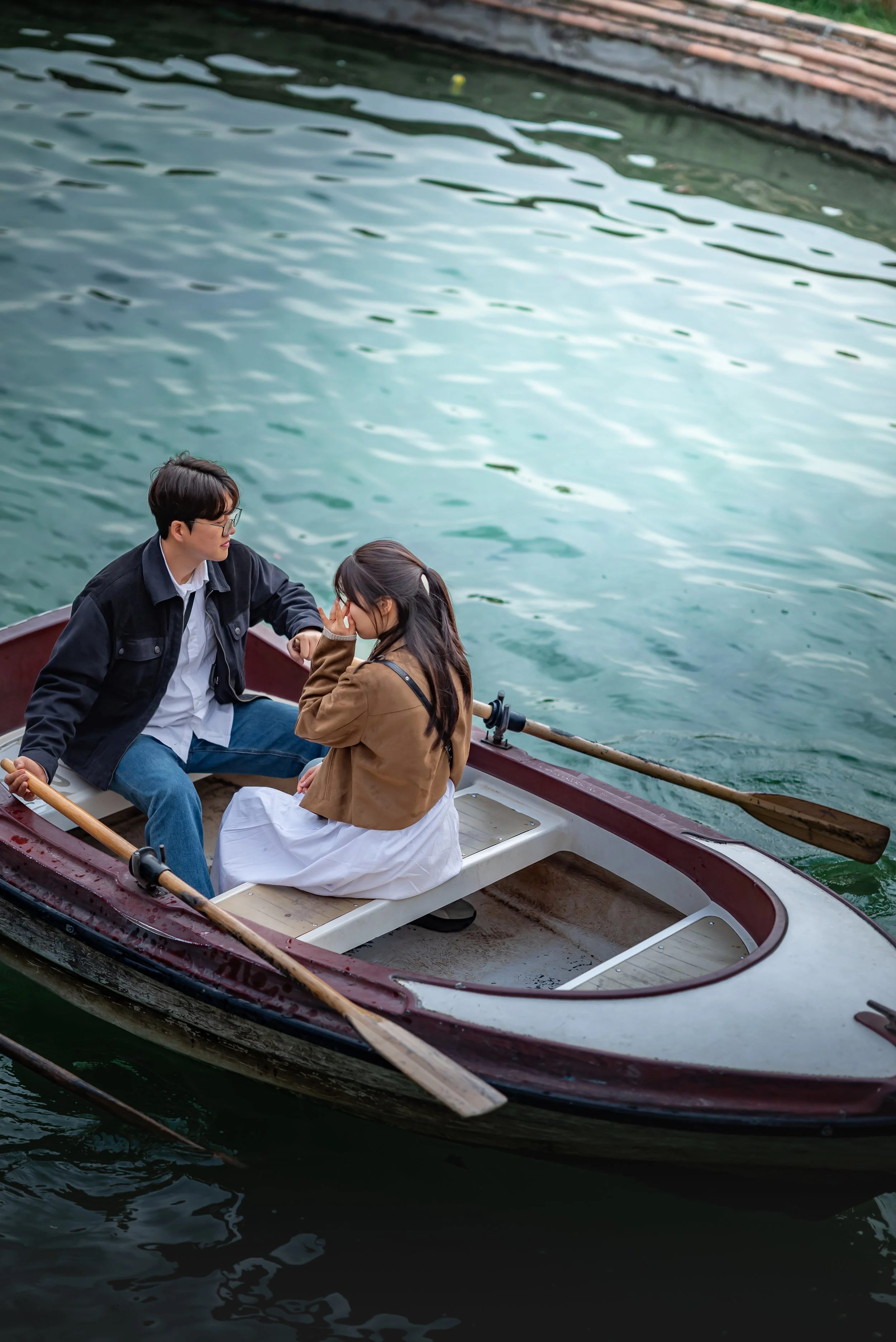 A man and woman sitting in a small rowboat on a body of water with ripples, with a wooden dock in the background. The man is wearing glasses, a black jacket, and blue jeans, while the woman is wearing a brown jacket and a white skirt. The woman is to